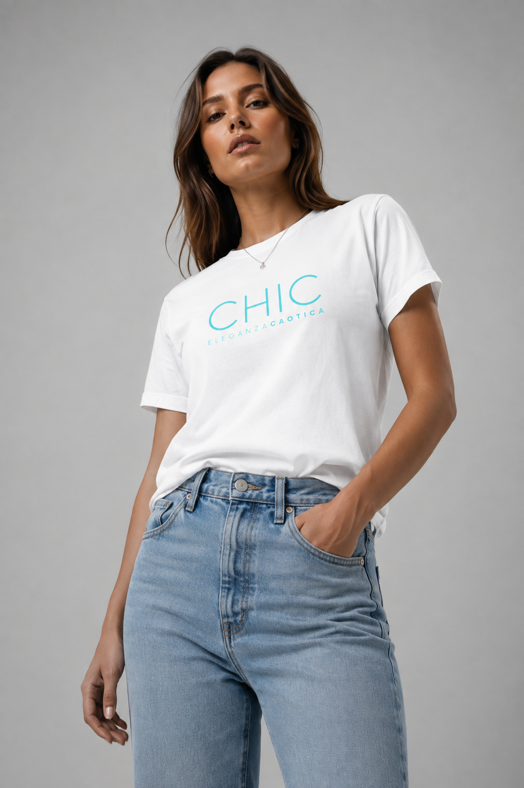 Woman wearing a white CHIC Eleganza Caotica graphic T-shirt with turquoise text, paired with light-wash blue jeans in a minimalist gray studio.
