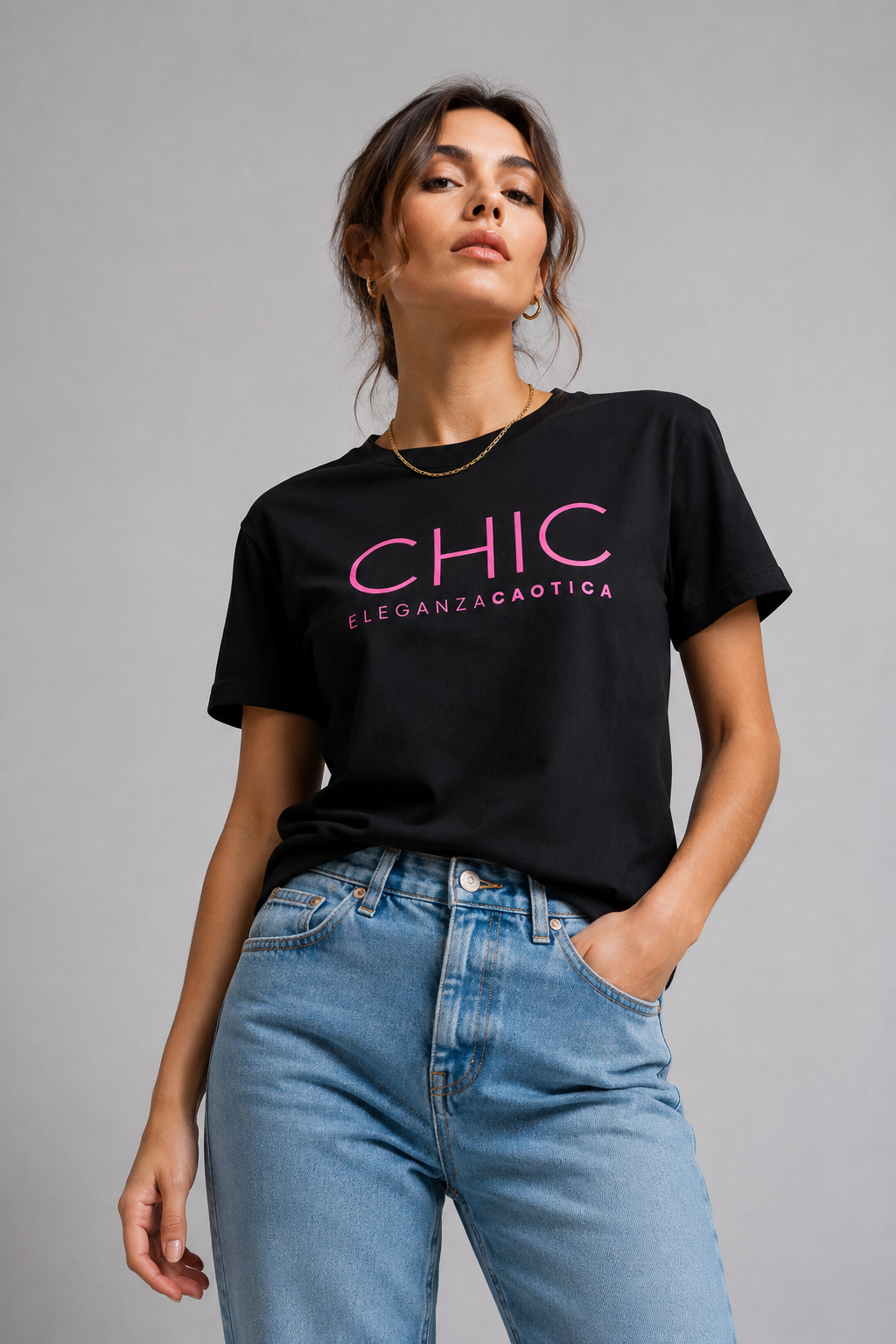 A woman wearing a black graphic T-shirt with pink text reading 'CHIC ELEGANZA CAOTICA' and light blue jeans, posing against a neutral gray studio background.