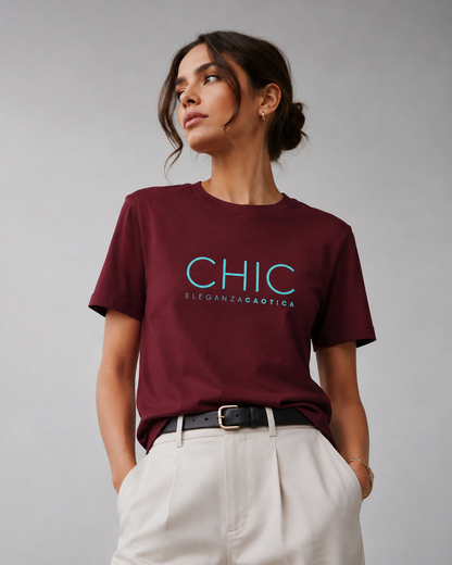 Woman wearing a maroon CHIC Eleganza Caotica graphic T-shirt with light blue typography, styled with cream trousers and a black belt in a neutral gray studio.