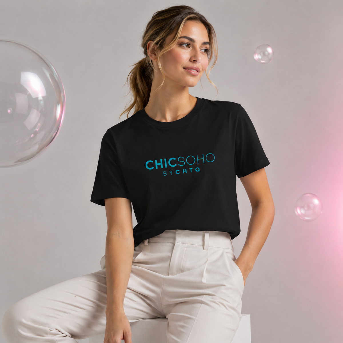 Woman wearing a black CHICSOHO graphic T-shirt with teal logo text and white trousers in a light gray editorial studio with floating bubbles.