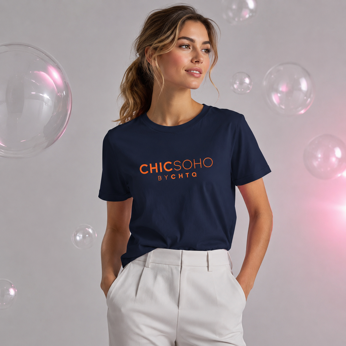 Woman wearing a navy blue CHICSOHO graphic T-shirt with orange logo text and white trousers in a light gray editorial studio with floating bubbles.