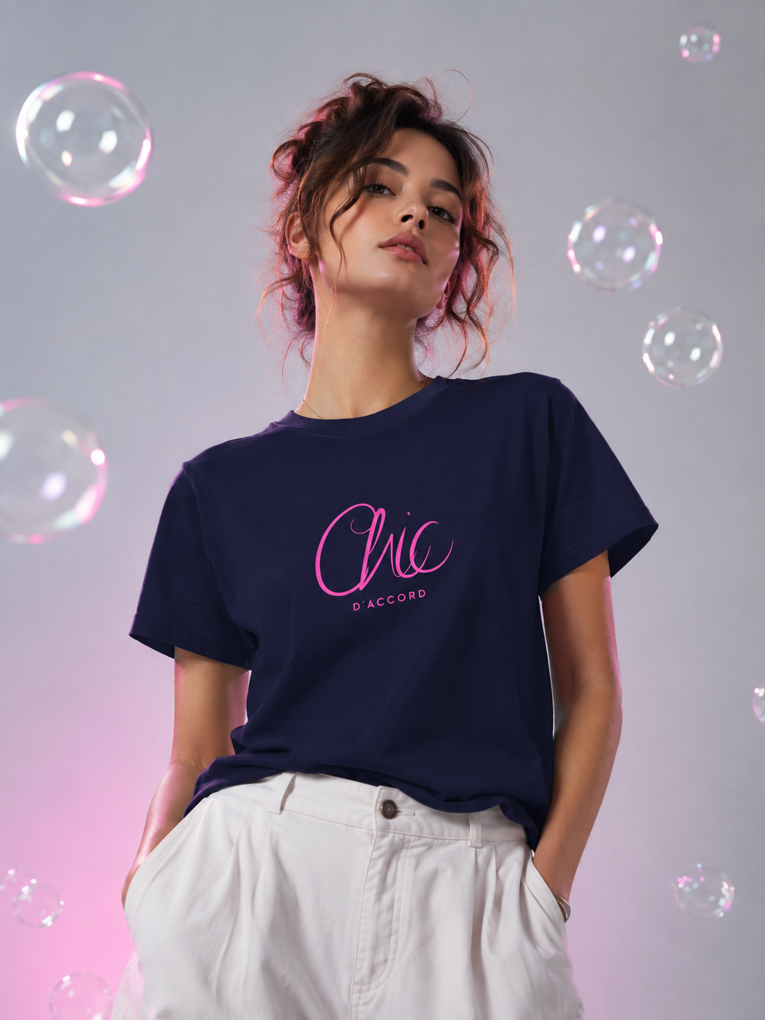 A woman wearing a navy blue t-shirt with a pink 'Chic D'Accord' graphic, paired with white pants, set against a gray background with floating bubbles.

