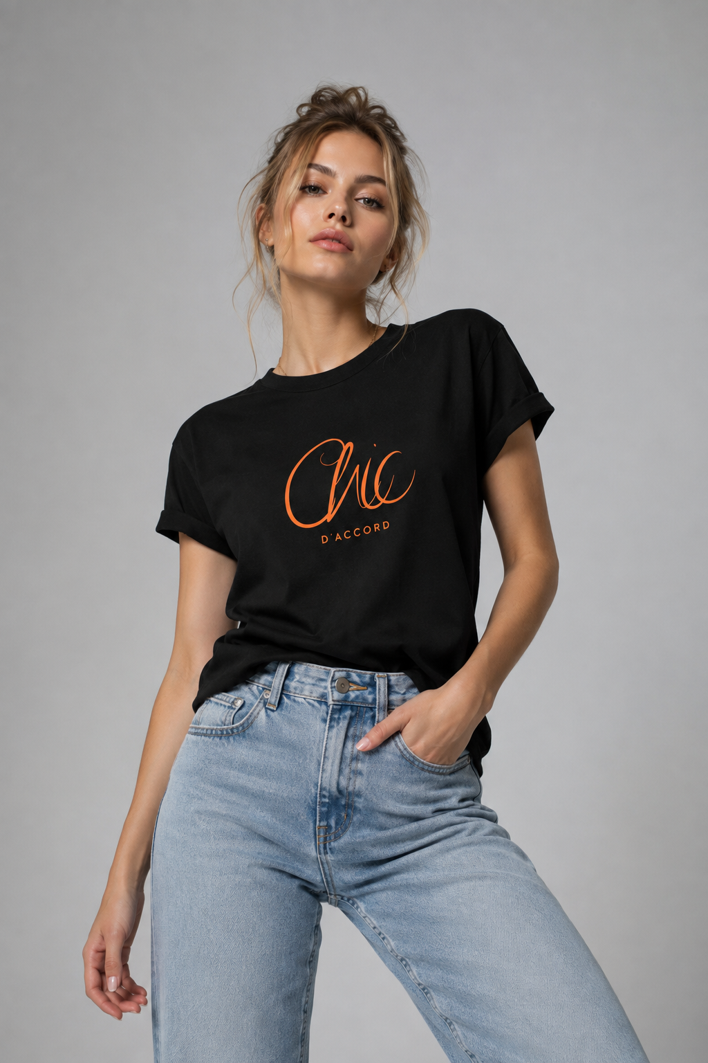 A woman modeling a black short-sleeve graphic T-shirt with orange cursive text reading 'Chic' and smaller text 'D'ACCORD' underneath, paired with light blue jeans against a plain gray studio background.