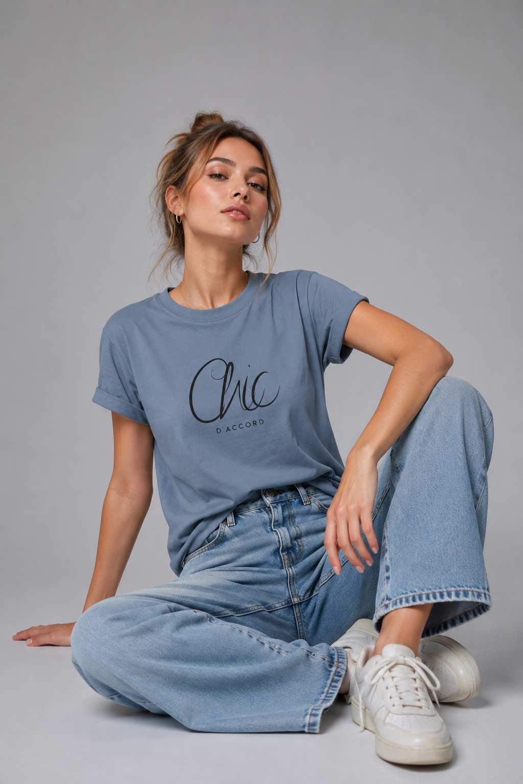 A woman sits cross-legged on a light gray studio floor wearing a dusty blue T-shirt with a black “Chic D’Accord” graphic, light-wash wide-leg jeans, and white sneakers.