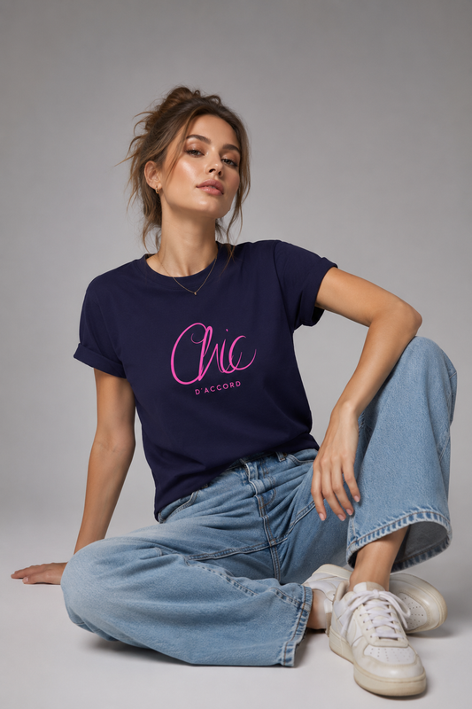 A woman sits cross-legged on a light gray studio floor wearing a navy blue T-shirt with a pink “Chic D’Accord” graphic, light-wash wide-leg jeans, and white sneakers.