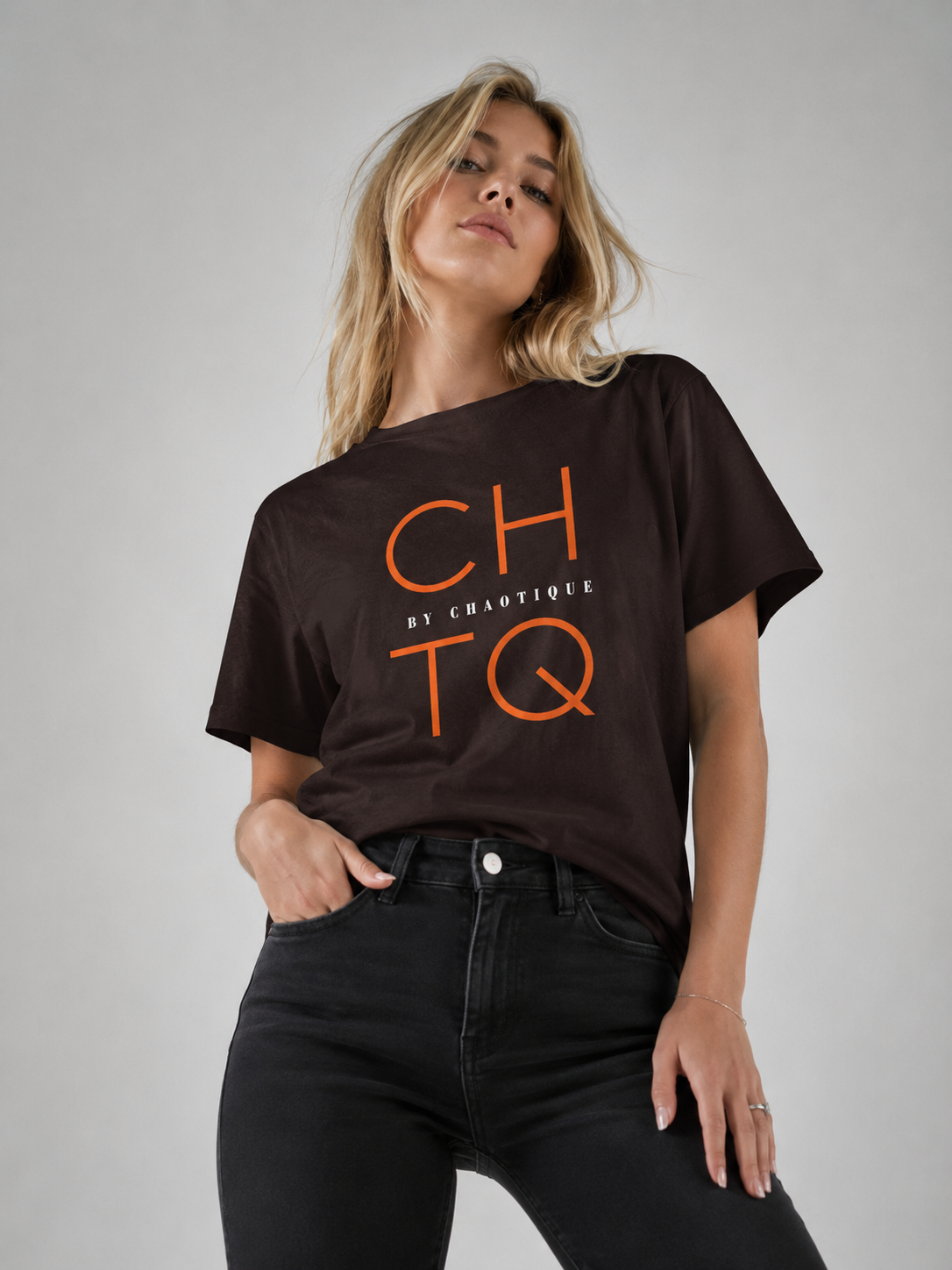 A woman wearing a dark brown short-sleeve graphic t-shirt with orange 'CHTQ' and 'BY CHAOTIQUE' text, paired with black jeans, against a neutral gray studio background.