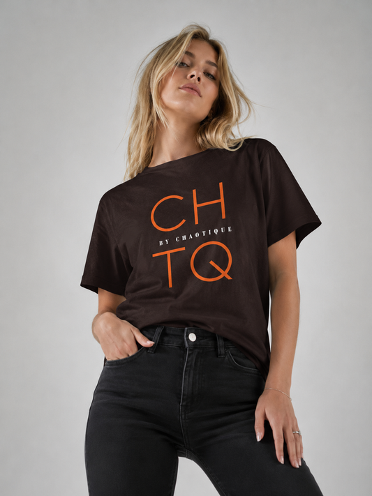 A woman wearing a dark brown short-sleeve graphic t-shirt with orange 'CHTQ' and 'BY CHAOTIQUE' text, paired with black jeans, against a neutral gray studio background.