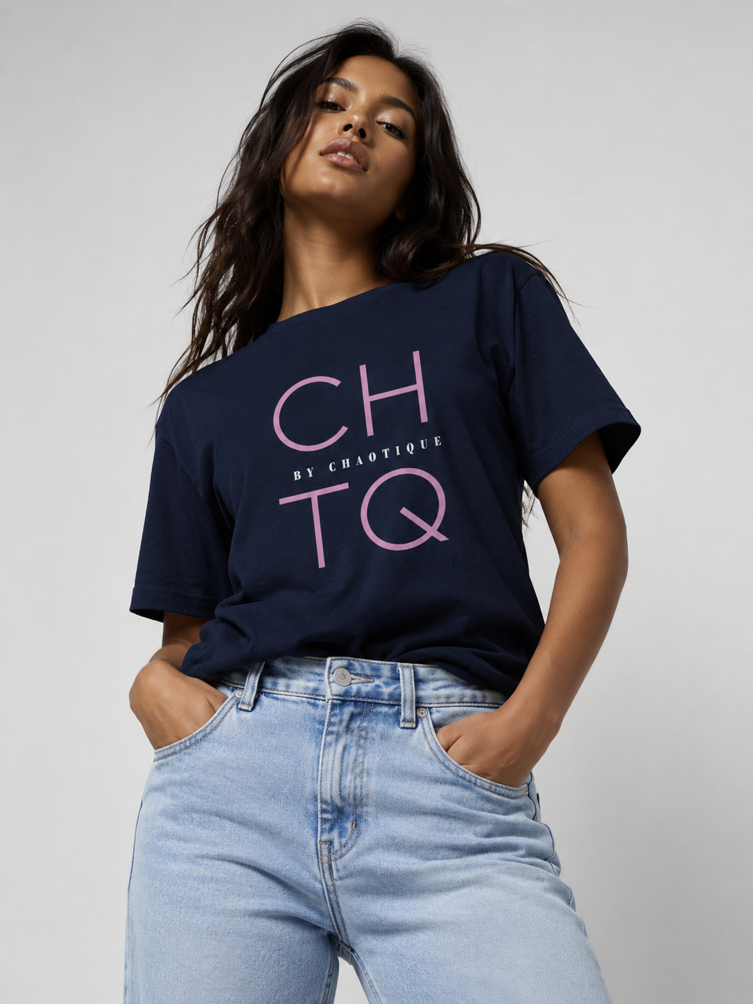 A woman wearing a navy blue off-the-shoulder graphic t-shirt with pink 'CHTQ' and 'By Chaotique' lettering and light wash blue jeans, posing against a neutral light gray background.