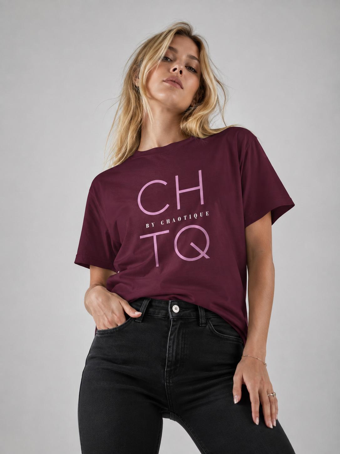 A woman wearing a maroon short-sleeve graphic t-shirt with 'CHTQ BY CHAOTIQUE' printed in light pink on the front, paired with black jeans, against a solid gray studio background.