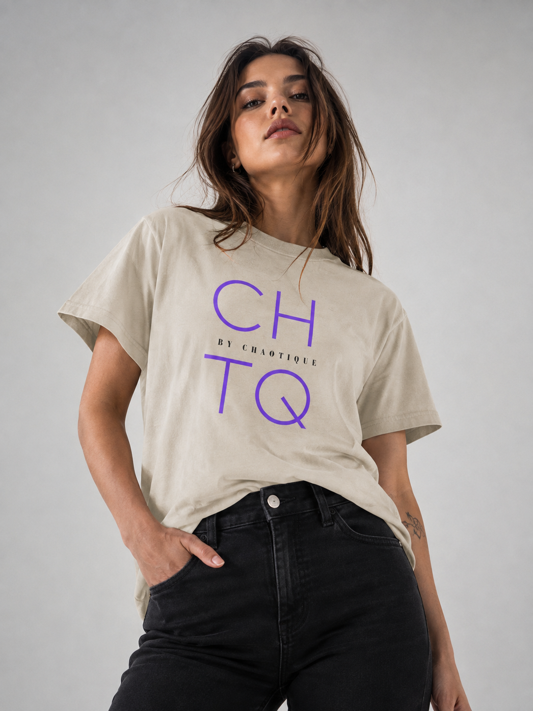 A woman wearing a beige graphic T-shirt with purple “CHTQ” typography and small “BY CHAOTIQUE” text on the front, paired with black jeans, posing against a neutral gray studio background.