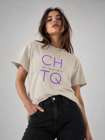 A woman wearing a beige graphic T-shirt with purple “CHTQ” typography and small “BY CHAOTIQUE” text on the front, paired with black jeans, posing against a neutral gray studio background.