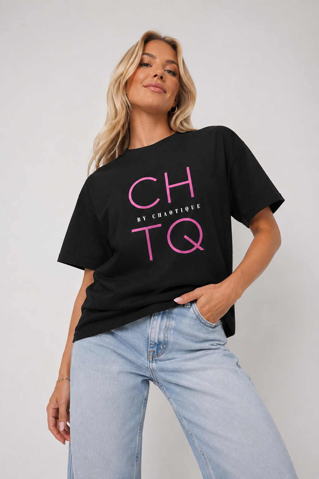A woman wearing a black oversized graphic T-shirt with pink 'CHTQ' lettering and 'BY CHAOTIQUE' text, paired with light blue jeans, against a plain light gray studio background.