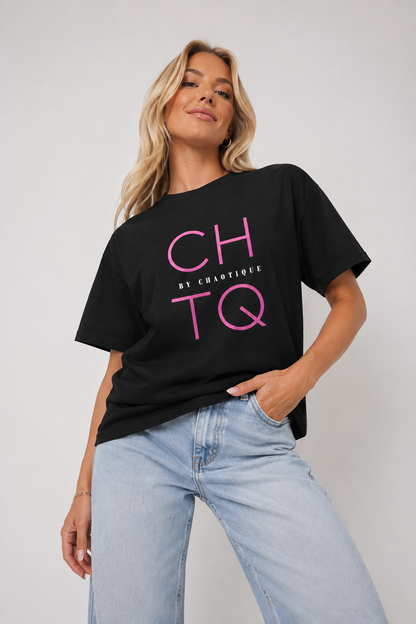 A woman wearing a black oversized graphic T-shirt with pink 'CHTQ' lettering and 'BY CHAOTIQUE' text, paired with light blue jeans, against a plain light gray studio background.
