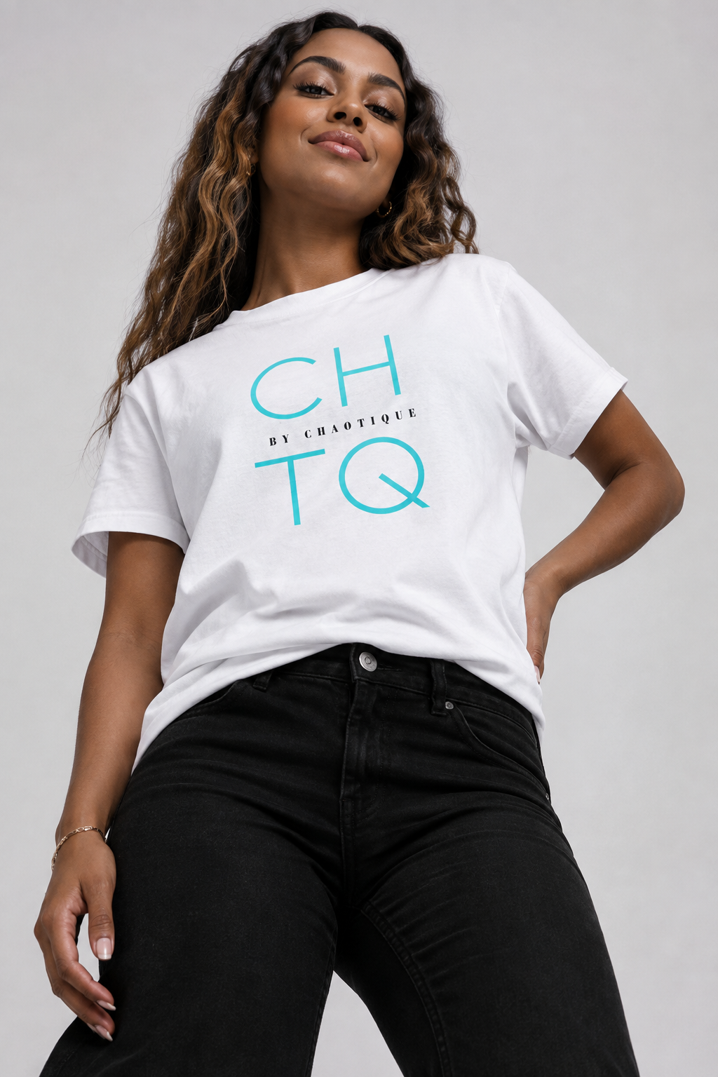 A woman wearing a white graphic t-shirt with 'CHTQ BY CHAOTIQUE' printed in teal, paired with black jeans, against a light gray studio background.