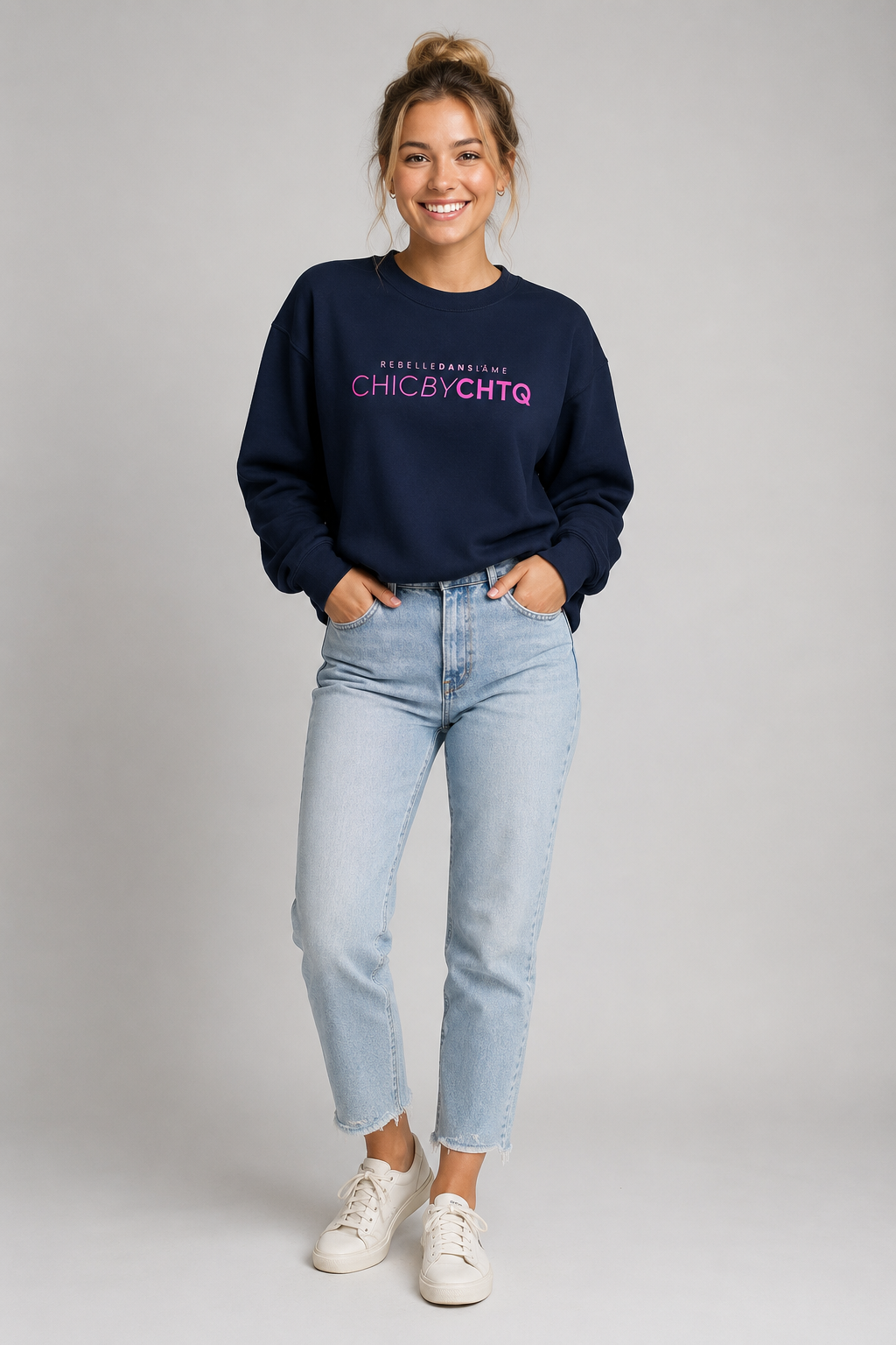 A woman modeling a navy blue crewneck sweatshirt with pink text reading “CHIC BY CHTQ” and “REBELLE DANS L’ÂME” above it, paired with light blue jeans and white sneakers against a light gray studio background.