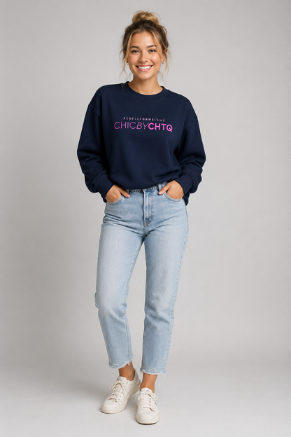 A woman modeling a navy blue crewneck sweatshirt with pink text reading “CHIC BY CHTQ” and “REBELLE DANS L’ÂME” above it, paired with light blue jeans and white sneakers against a light gray studio background.
