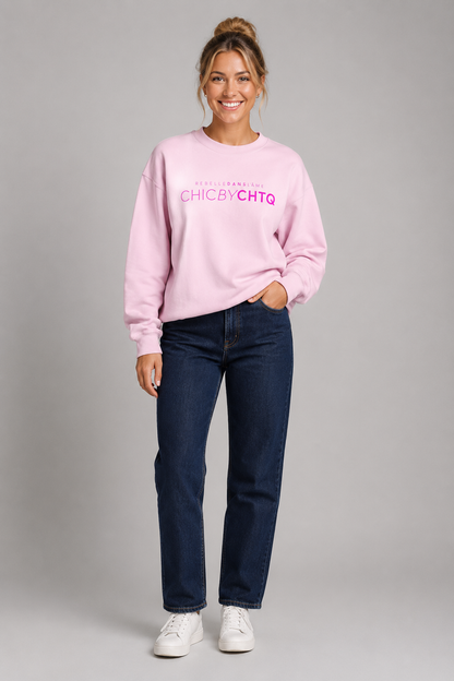 A woman modeling a lightpink crewneck sweatshirt with pink text reading “CHIC BY CHTQ” and “REBELLE DANS L’ÂME” above it, paired with light blue jeans and white sneakers against a light gray studio background.