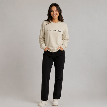 A woman modeling a beige crewneck sweatshirt with dark grey text reading “CHIC BY CHTQ” and “REBELLE DANS L’ÂME” above it, paired with light blue jeans and white sneakers against a light gray studio background.