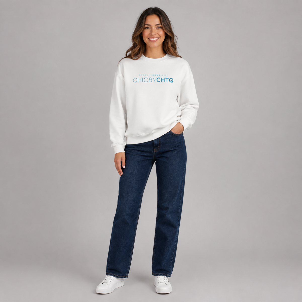 A woman modeling a white crewneck sweatshirt with turquoise text reading “CHIC BY CHTQ” and “REBELLE DANS L’ÂME” above it, paired with light blue jeans and white sneakers against a light gray studio background.