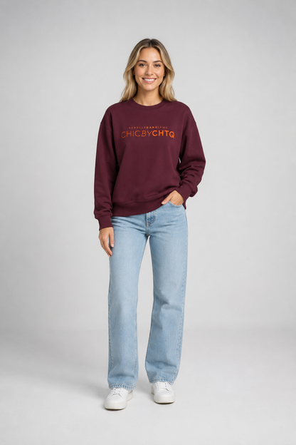 A woman modeling a maroon crewneck sweatshirt with orange text reading “CHIC BY CHTQ” and “REBELLE DANS L’ÂME” above it, paired with light blue jeans and white sneakers against a light gray studio background.