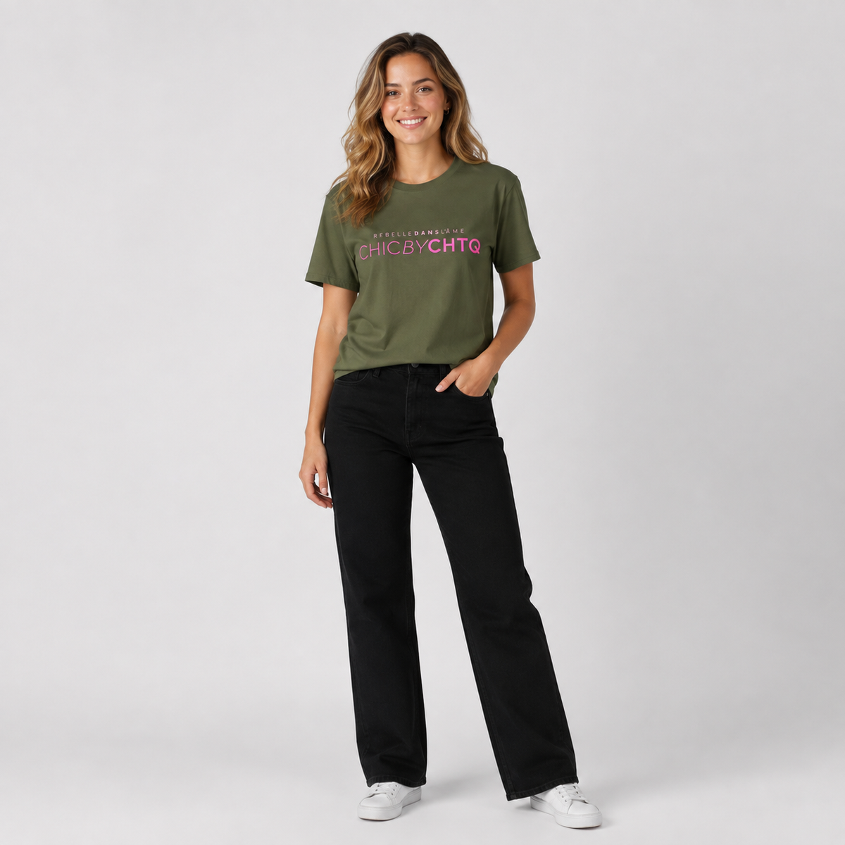 A woman modeling an olive green graphic T-shirt featuring pink “CHIC BY CHTQ” text with “REBELLE DANS L’ÂME” above, paired with black wide-leg jeans and white sneakers against a cool light gray studio background.