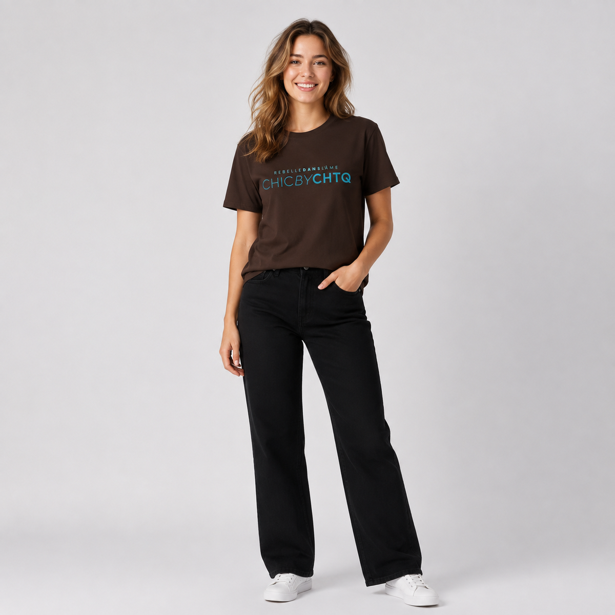 A woman modeling a dark brown short-sleeve graphic T-shirt featuring light blue and white “CHIC BY CHTQ” text with “REBELLE DANS L’ÂME” above, paired with black wide-leg jeans and white sneakers against a cool light gray studio background.