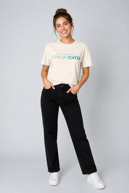 A woman modeling a cream-colored graphic T-shirt featuring turquoise “CHIC BY CHTQ” text with “REBELLE DANS L’ÂME” above, paired with black jeans and white sneakers against a cool light gray studio background.