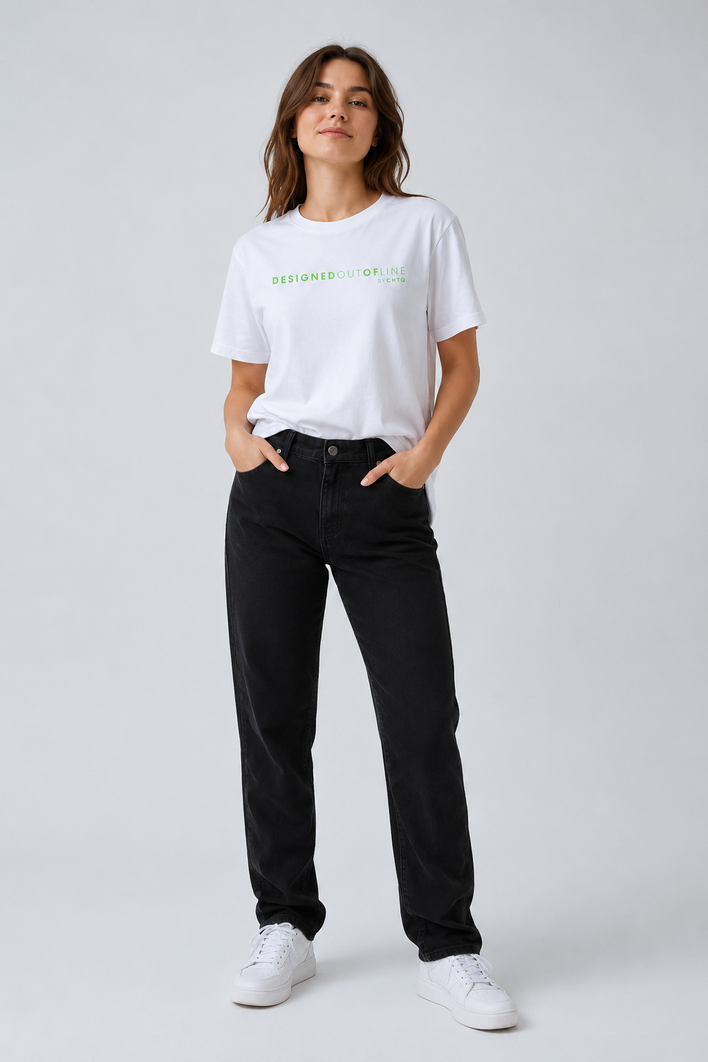 A woman modeling a white crewneck T-shirt with green text that reads “DESIGNED OUT OF LINE” and “BY CHTQ” across the chest, paired with black jeans and white sneakers, against a light gray studio background.