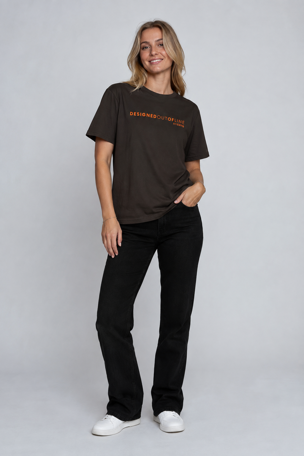 A woman modeling a dark brown short-sleeve T-shirt with orange text reading “DESIGNED OUT OF LINE” and “BY CHTQ” on the chest, paired with black jeans and white sneakers, against a cool light gray studio background.