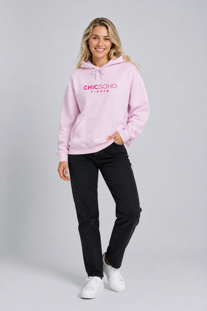 A woman modeling a light pink pullover hoodie with “CHICSOHO BY CHTQ” printed in pink across the chest, paired with black pants and white sneakers, against a light gray studio background.