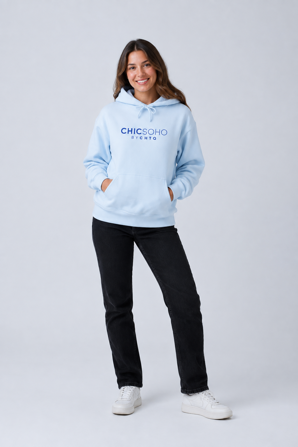 A woman modeling a light blue pullover hoodie with “CHICSOHO BY CHTQ” printed in blue across the chest, paired with black pants and white sneakers, against a light gray studio background.