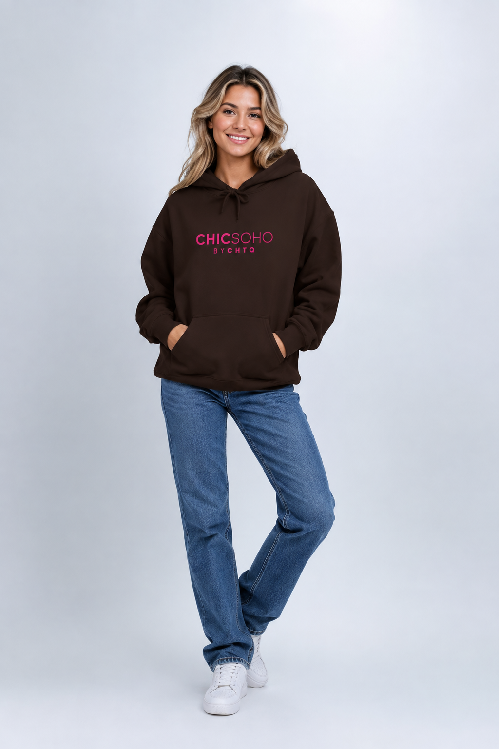 A woman modeling a brown pullover hoodie with “CHICSOHO BY CHTQ” printed in pink across the chest, paired with black pants and white sneakers, against a light gray studio background.