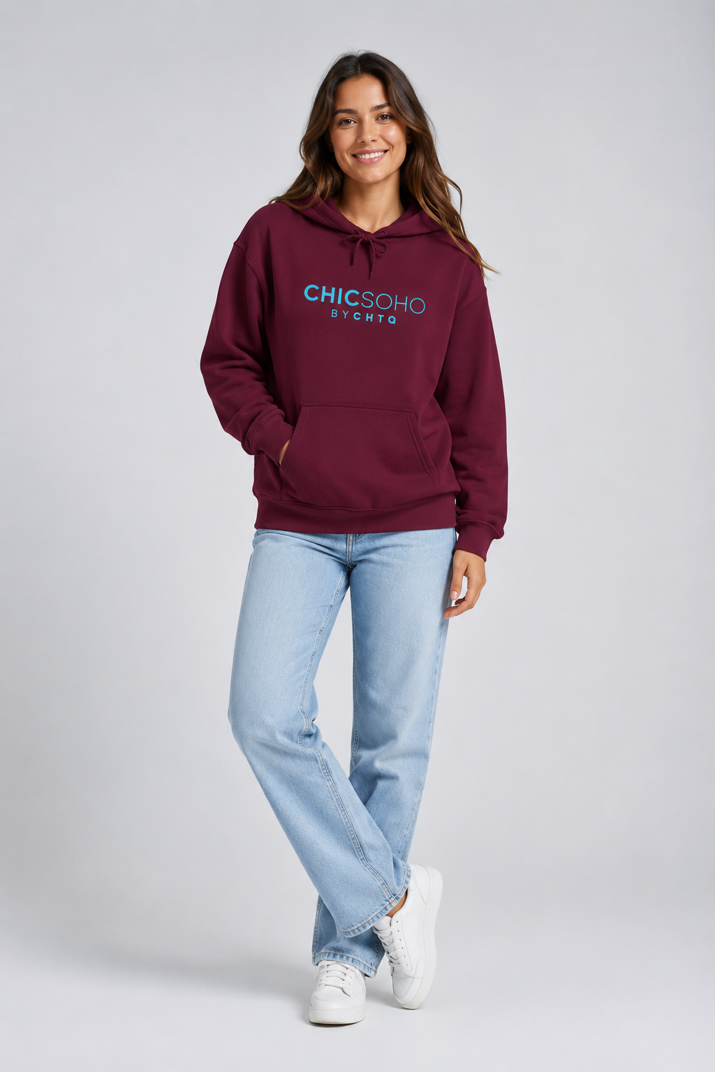 A woman modeling a maroon pullover hoodie with “CHICSOHO BY CHTQ” printed in turquoise across the chest, paired with black pants and white sneakers, against a light gray studio background.