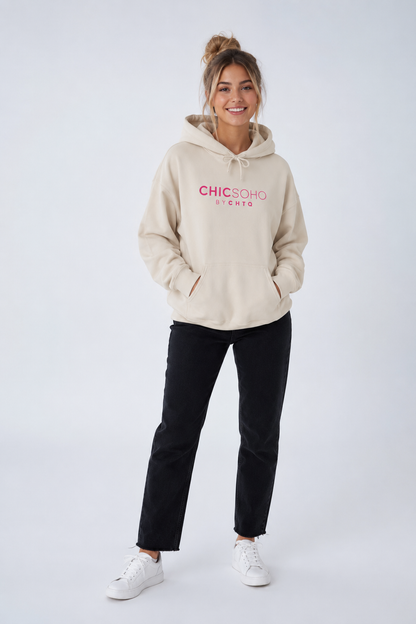 A woman modeling a beige pullover hoodie with “CHICSOHO BY CHTQ” printed in pink across the chest, paired with black pants and white sneakers, against a light gray studio background.
