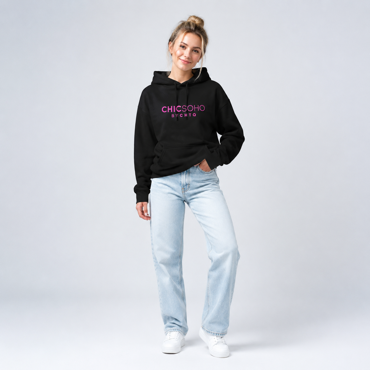 A woman modeling a black pullover hoodie with “CHICSOHO BY CHTQ” printed in pink across the chest, paired with black pants and white sneakers, against a light gray studio background.