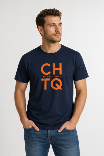 A man wearing a navy blue graphic T-shirt with “CHTQ BY CHAOTIQUE” printed on the chest, paired with blue jeans and white sneakers, standing against a light gray studio background.