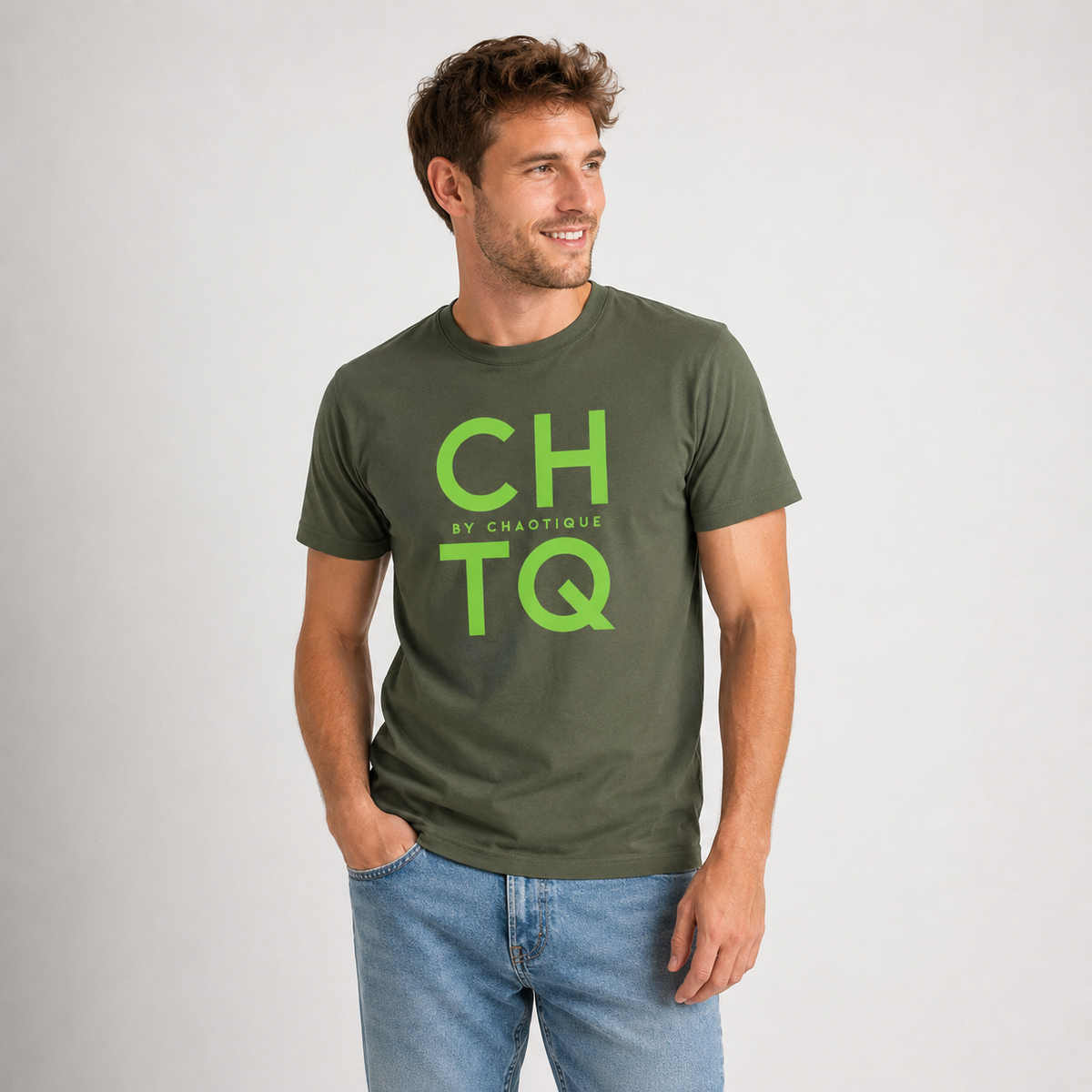 A man wearing a green graphic T-shirt with “CHTQ BY CHAOTIQUE” printed on the chest, paired with blue jeans and white sneakers, standing against a light gray studio background.