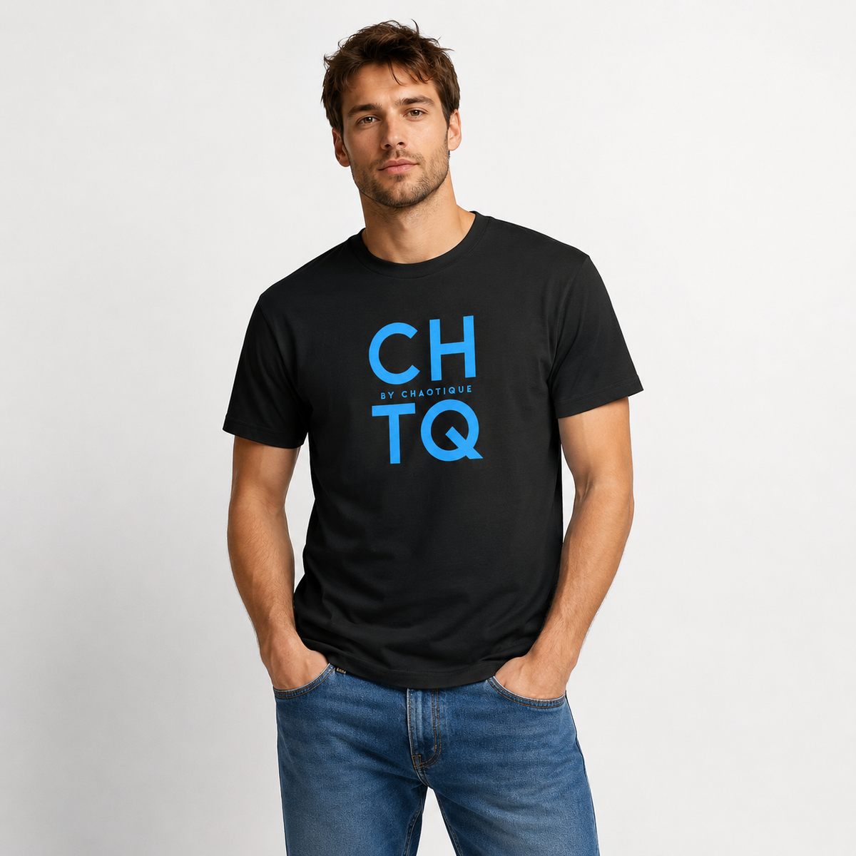 A man wearing a black graphic T-shirt with “CHTQ BY CHAOTIQUE” printed on the chest, paired with blue jeans and white sneakers, standing against a light gray studio background.