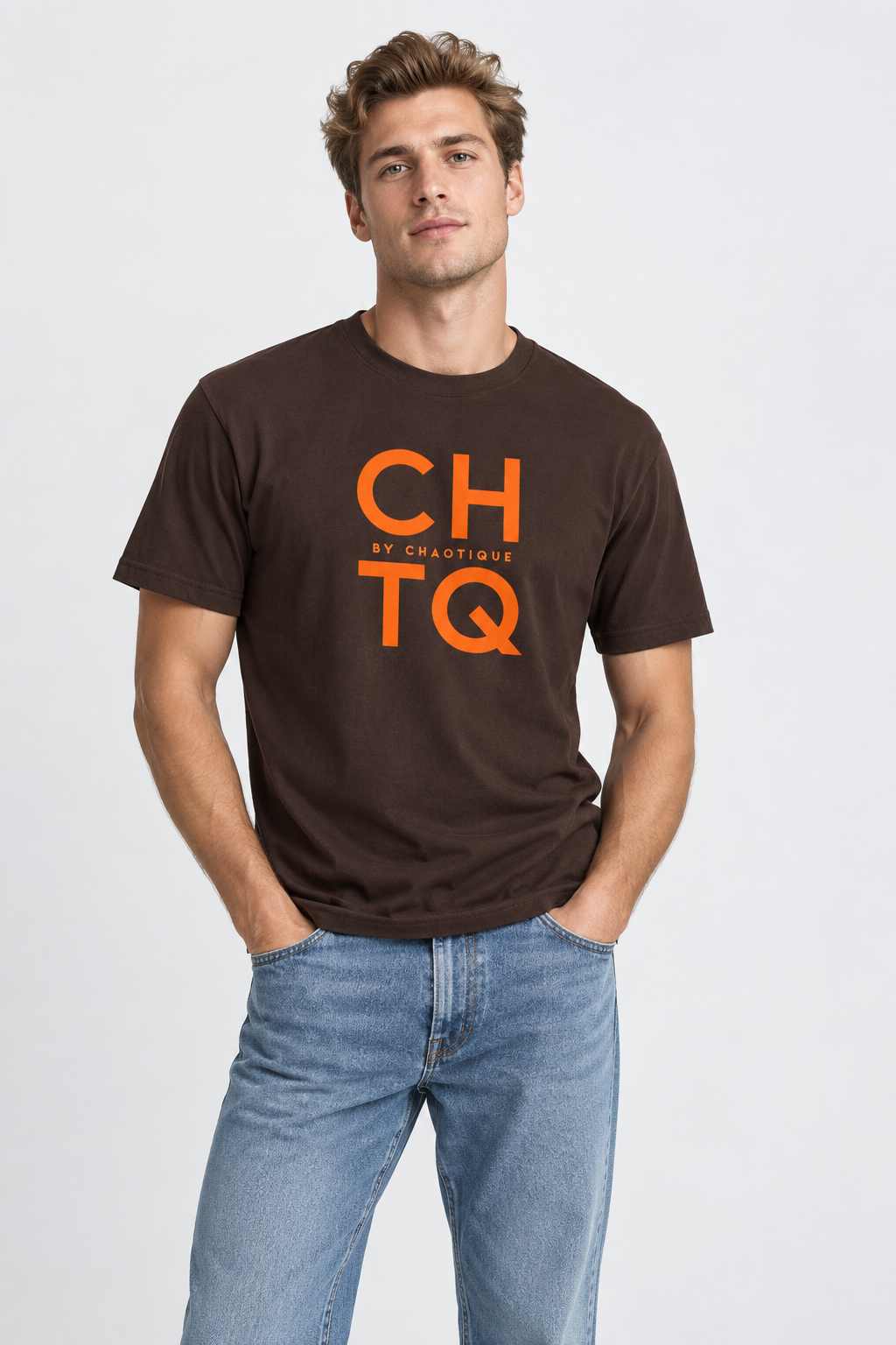 A man wearing a brown graphic T-shirt with “CHTQ BY CHAOTIQUE” printed on the chest, paired with blue jeans and white sneakers, standing against a light gray studio background.