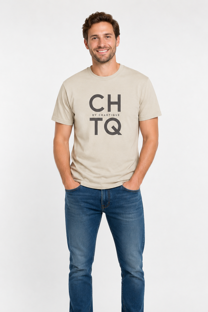 A man wearing a beige graphic T-shirt with “CHTQ BY CHAOTIQUE” printed on the chest, paired with blue jeans and white sneakers, standing against a light gray studio background.