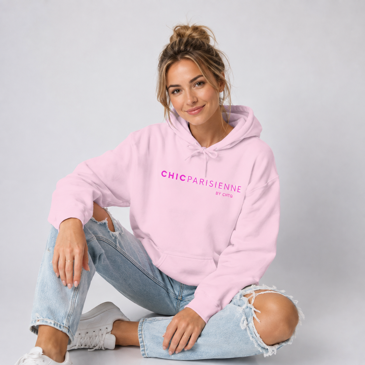 A woman sitting cross-legged on a light gray studio floor, wearing a light pink hoodie with pink “CHIC PARISIENNE BY CHTQ” text printed across the chest, paired with light-wash ripped jeans and white sneakers.