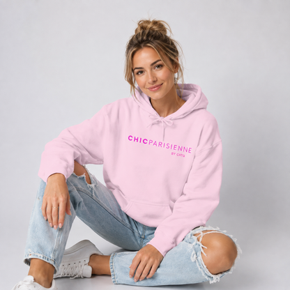 A woman sitting cross-legged on a light gray studio floor, wearing a light pink hoodie with pink “CHIC PARISIENNE BY CHTQ” text printed across the chest, paired with light-wash ripped jeans and white sneakers.