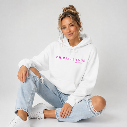 A woman sitting cross-legged on a light gray studio floor, wearing a white hoodie with pink “CHIC PARISIENNE BY CHTQ” text printed across the chest, paired with light-wash ripped jeans and white sneakers.