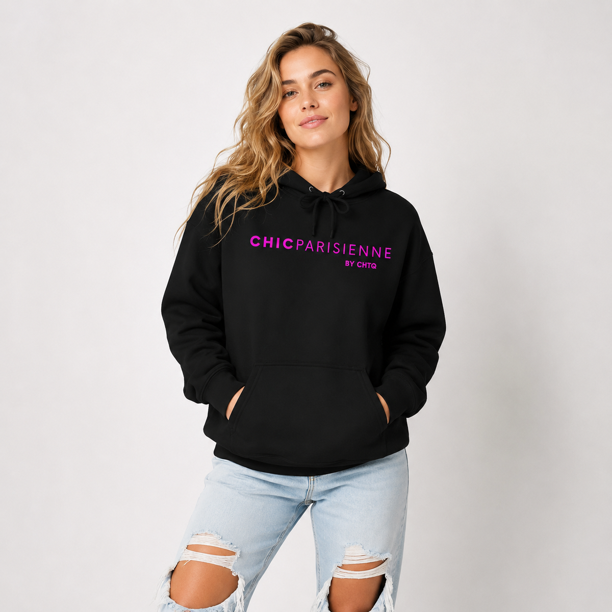 Woman wearing a black CHIC PARISIENNE BY CHTQ graphic hoodie with bright pink lettering, paired with light-wash ripped jeans in a light studio setting.