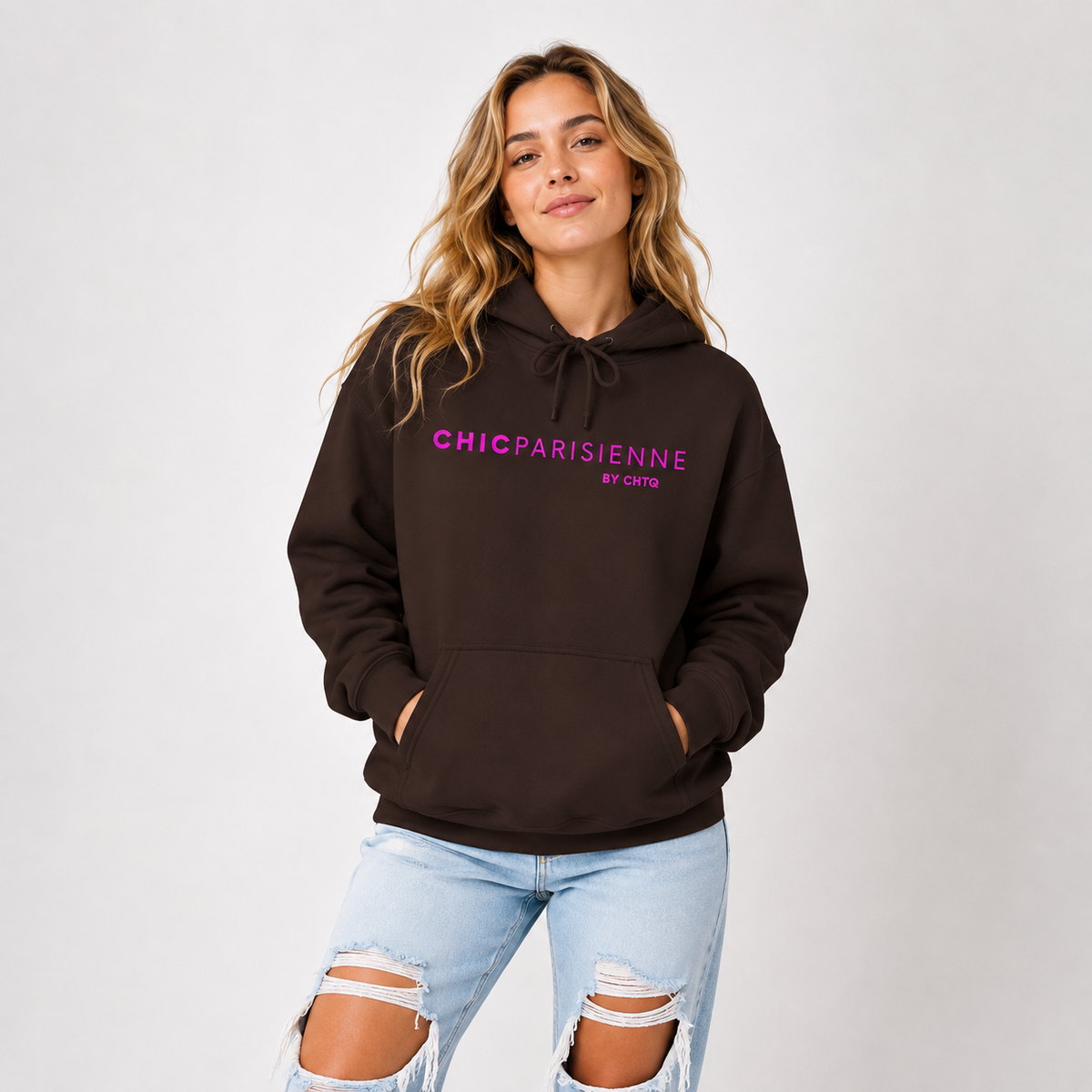 Woman wearing a brown CHIC PARISIENNE BY CHTQ graphic hoodie with bright pink lettering, paired with light-wash ripped jeans in a light studio setting.