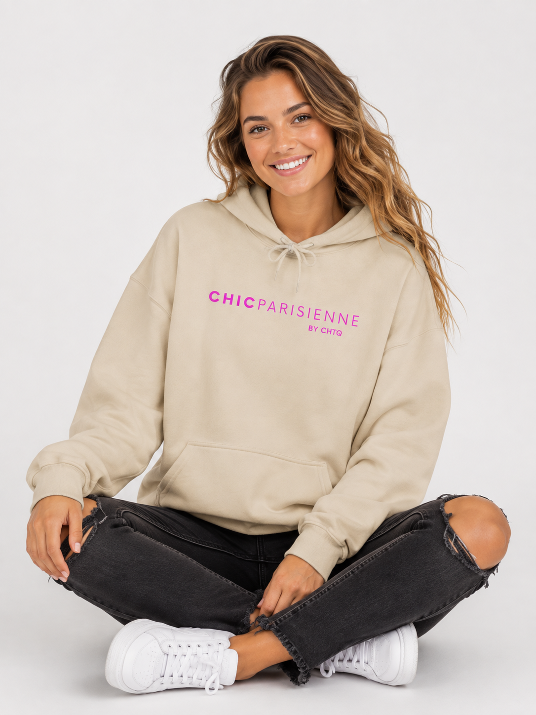 A woman sitting cross-legged on a light gray studio floor, wearing a beige hoodie with pink “CHIC PARISIENNE BY CHTQ” text printed across the chest, paired with black ripped jeans and white sneakers.