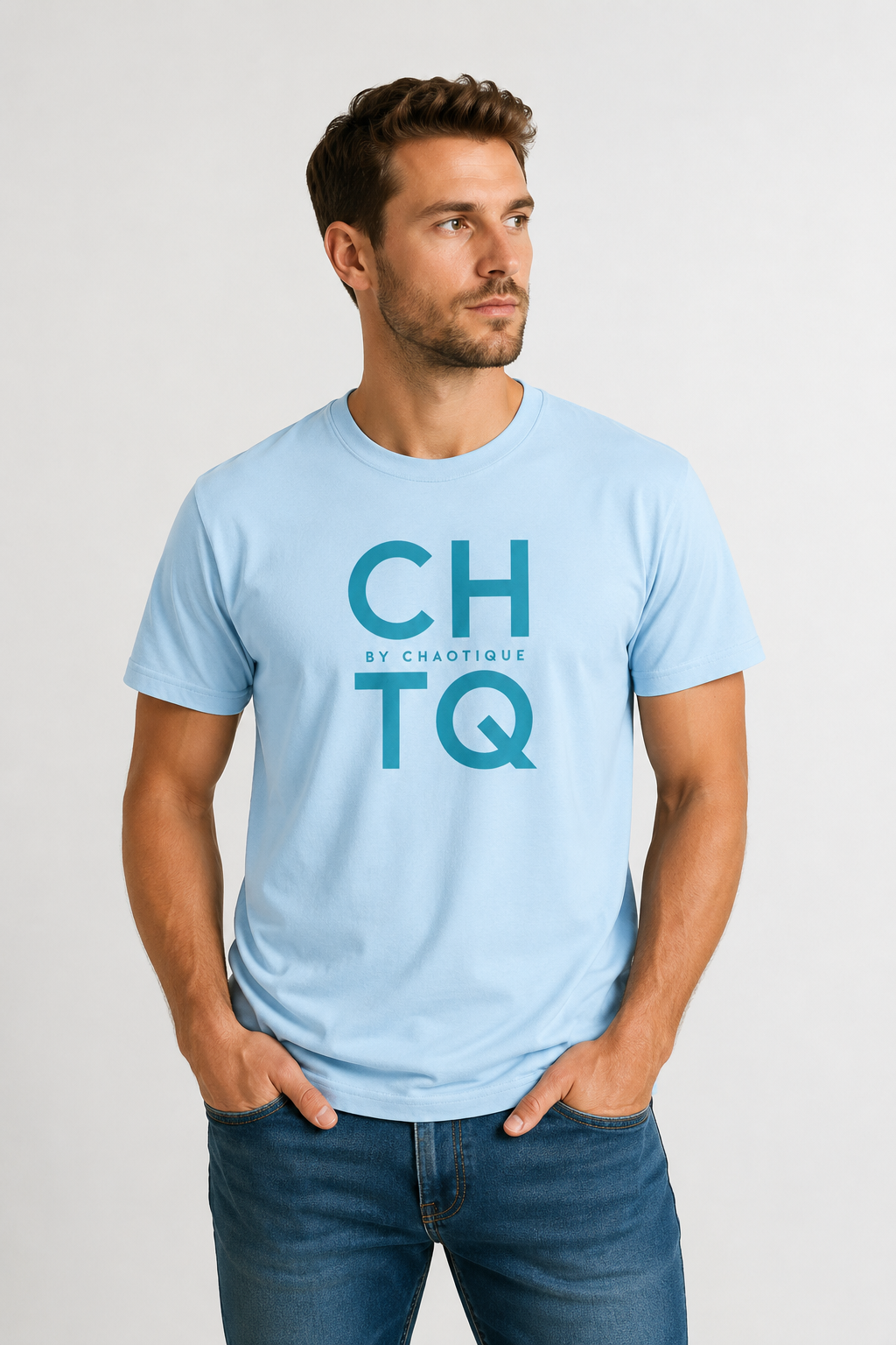 A man wearing a light blue graphic T-shirt with “CHTQ BY CHAOTIQUE” printed on the chest, paired with blue jeans and white sneakers, standing against a light gray studio background.