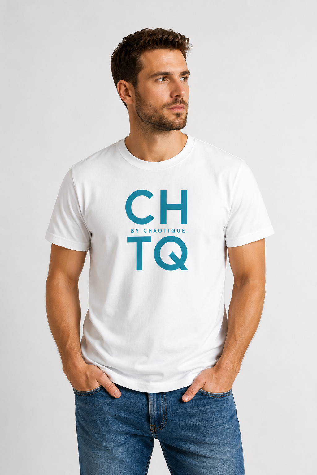 A man wearing a white graphic T-shirt with “CHTQ BY CHAOTIQUE” printed on the chest, paired with blue jeans and white sneakers, standing against a light gray studio background.