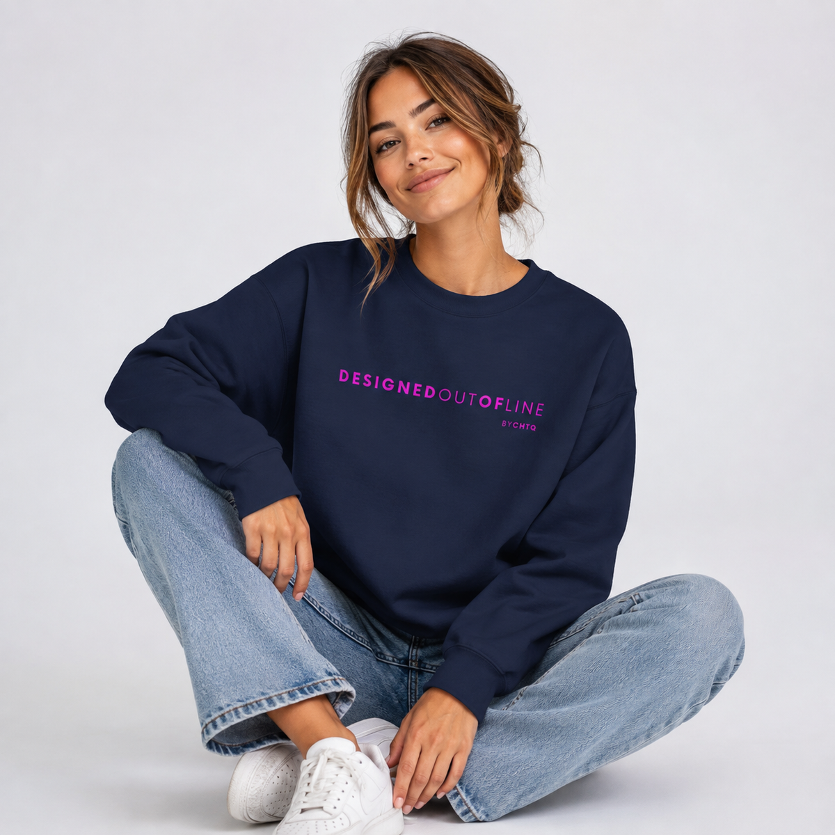 A woman sitting cross-legged against a light gray studio background, wearing a navy crewneck sweatshirt featuring pink “DESIGNED OUT OF LINE” text across the chest, paired with light blue jeans.