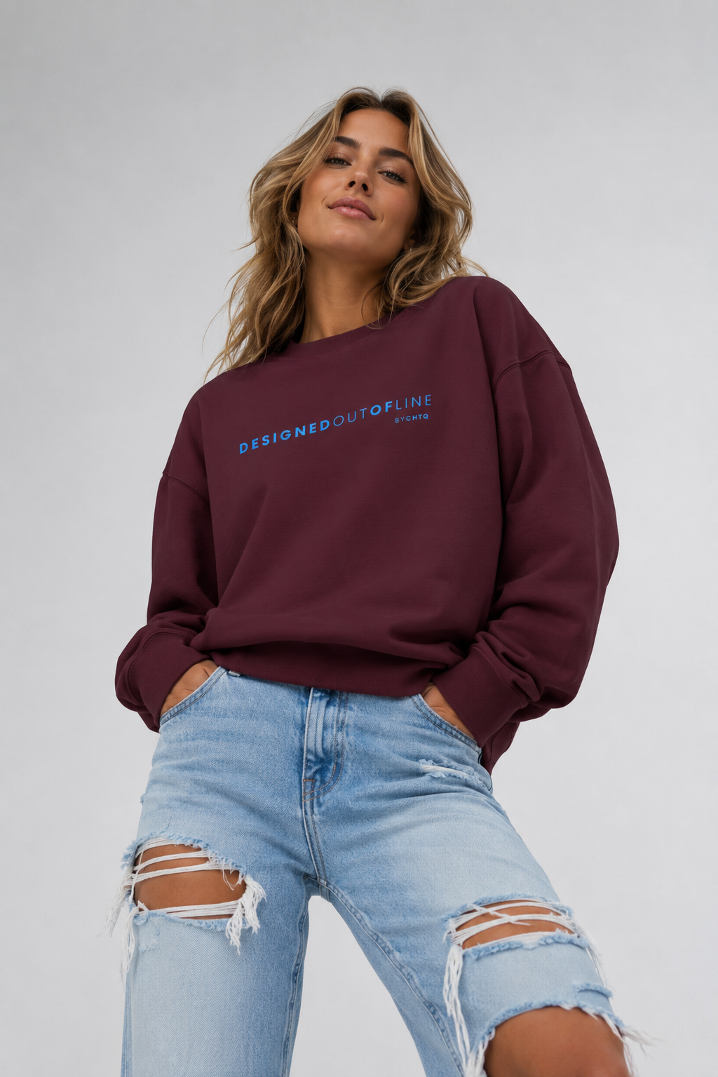 A woman wearing a maroon crewneck sweatshirt with “DESIGNED OUT OF LINE” printed in blue across the chest, paired with light-wash ripped jeans, photographed against a neutral light gray studio background.